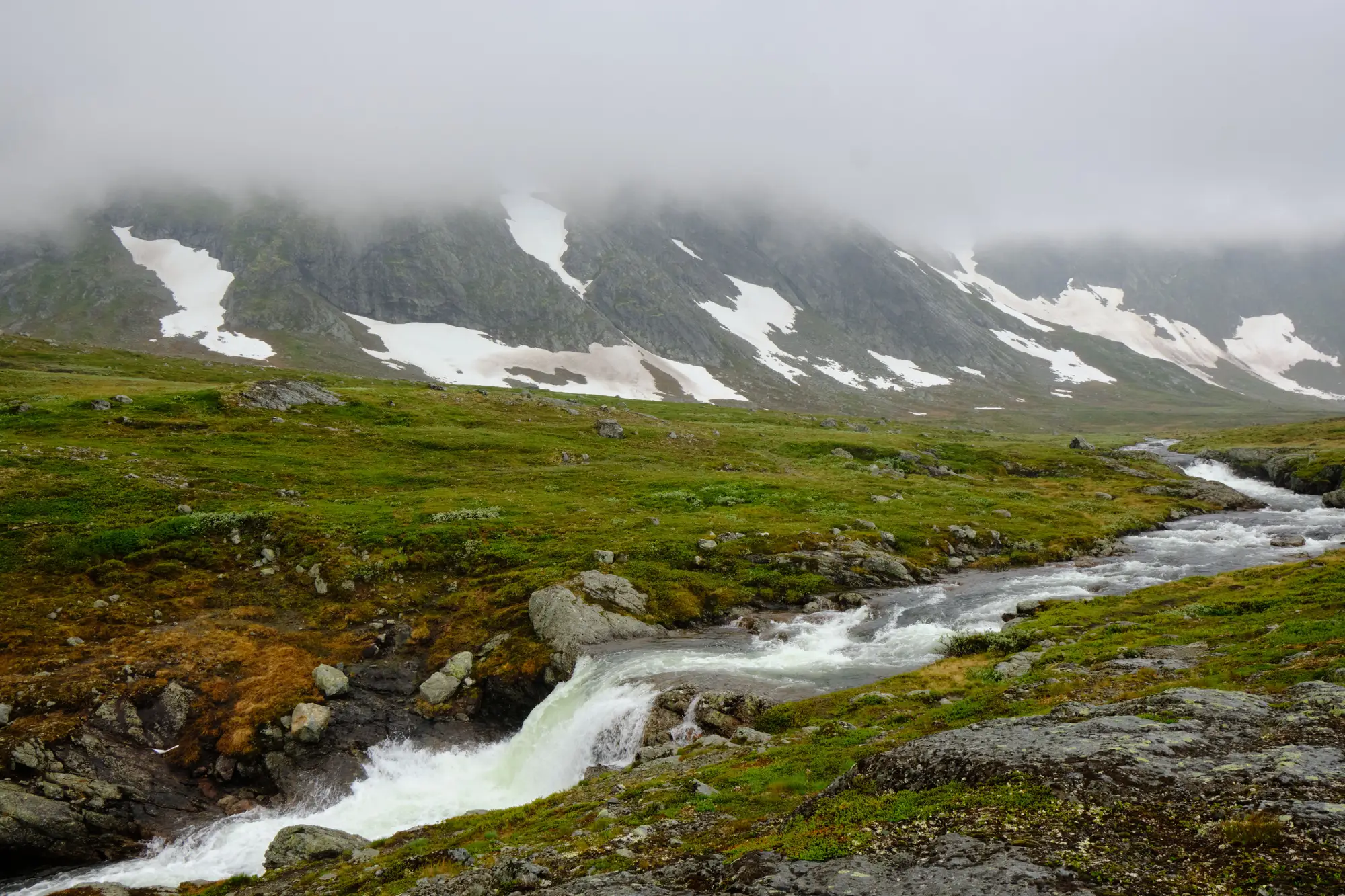 A meltwater stream running full below a mountainside where the snow has broken into patches, under low cloud